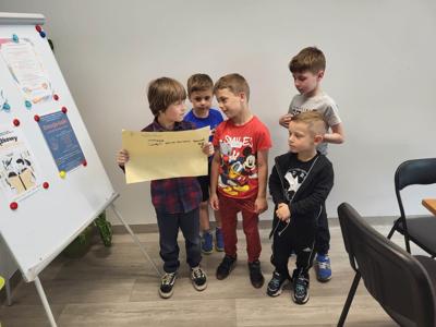 Na zdjęciu znajdują się uczestnicy warsztatów. Dzieci stoją i trzymają rękach kartkę. Zdjęcie wykonano w bibliotece.