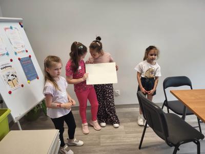 Na zdjęciu znajdują się uczestnicy warsztatów. Dzieci stoją i trzymają rękach kartkę. Zdjęcie wykonano w bibliotece.