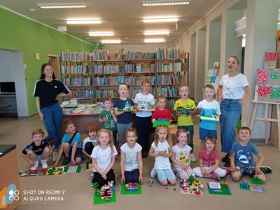 Na zdjęciu znajdują się uczestnicy warsztatów. Dzieci trzymają w ręce konstrukcje z klocków lego.  Zdjęcie wykonano w bibliotece.
