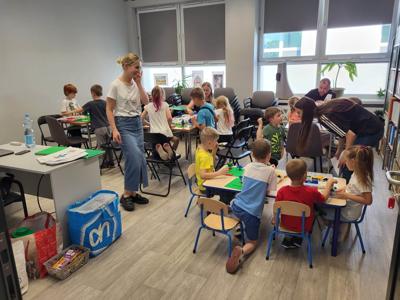 Na zdjęciu znajdują się uczestnicy warsztatów. Dzieci wykonają konstrukcje z klocków lego - siedzą na krzesłach. Zdjęcie wykonano w bibliotece.