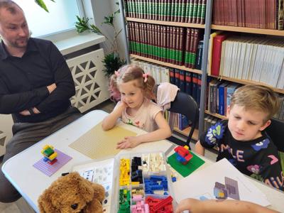 Na zdjęciu znajdują się uczestnicy warsztatów. Dzieci wykonają konstrukcje z klocków lego - siedzą na krzesłach. Zdjęcie wykonano w bibliotece.