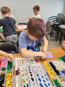 Na zdjęciu znajdują się uczestnicy warsztatów. Dzieci wykonają konstrukcje z klocków lego - siedzą na krzesłach. Zdjęcie wykonano w bibliotece.