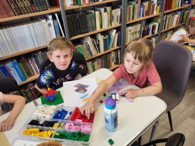 Na zdjęciu znajdują się uczestnicy warsztatów. Dzieci wykonają konstrukcje z klocków lego - siedzą na krzesłach. Zdjęcie wykonano w bibliotece.
