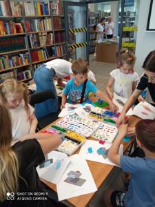 Na zdjęciu znajdują się uczestnicy warsztatów. Dzieci wykonają konstrukcje z klocków lego - siedzą na krzesłach. Zdjęcie wykonano w bibliotece.