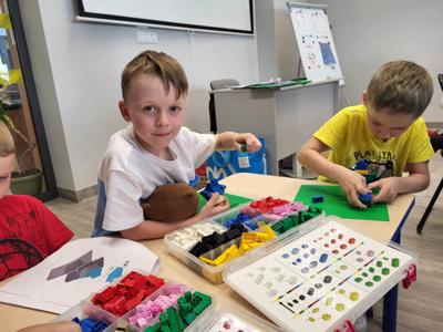 Na zdjęciu znajdują się uczestnicy warsztatów. Dzieci wykonają konstrukcje z klocków lego - siedzą na krzesłach. Zdjęcie wykonano w bibliotece.