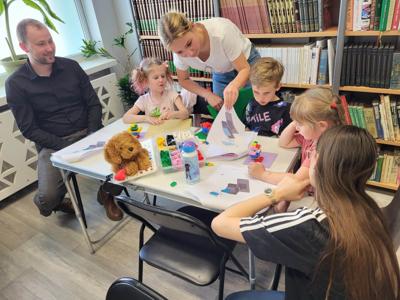 Na zdjęciu znajdują się uczestnicy warsztatów. Dzieci wykonają konstrukcje z klocków lego - siedzą na krzesłach. Zdjęcie wykonano w bibliotece.