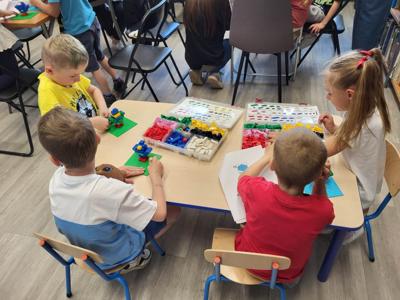 Na zdjęciu znajdują się uczestnicy warsztatów. Dzieci wykonają konstrukcje z klocków lego - siedzą na krzesłach. Zdjęcie wykonano w bibliotece.