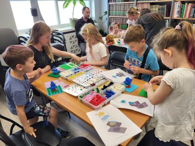 Na zdjęciu znajdują się uczestnicy warsztatów. Dzieci wykonają konstrukcje z klocków lego - siedzą na krzesłach. Zdjęcie wykonano w bibliotece.