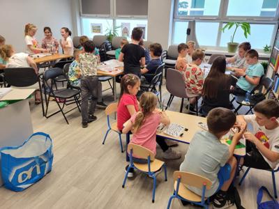 Na zdjęciu znajdują się uczestnicy warsztatów. Dzieci wykonają konstrukcje z klocków lego - siedzą na krzesłach. Zdjęcie wykonano w bibliotece.