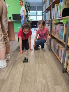 Na zdjęciu znajdują się uczestnicy warsztatów. Dzieci obserwują puszczane na podłodze samochody z klocków. Zdjęcie wykonano w bibliotece.