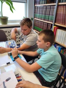 Na zdjęciu znajdują się uczestnicy warsztatów. Dzieci wykonają konstrukcje z klocków lego - siedzą na krzesłach. Zdjęcie wykonano w bibliotece.