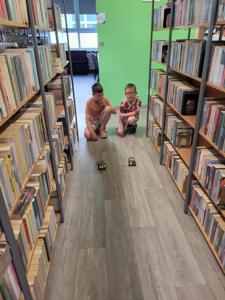 Na zdjęciu znajdują się uczestnicy warsztatów. Dzieci obserwują puszczane na podłodze samochody z klocków. Zdjęcie wykonano w bibliotece.