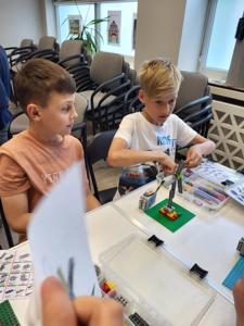 Na zdjęciu znajdują się uczestnicy warsztatów. Dzieci wykonają konstrukcje z klocków lego - siedzą na krzesłach. Zdjęcie wykonano w bibliotece.