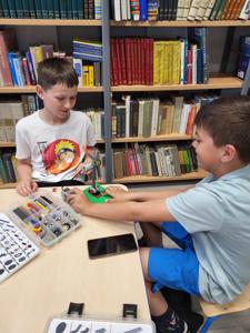 Na zdjęciu znajdują się uczestnicy warsztatów. Dzieci wykonają konstrukcje z klocków lego - siedzą na krzesłach. Zdjęcie wykonano w bibliotece.