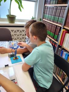 Na zdjęciu znajdują się uczestnicy warsztatów. Dzieci wykonają konstrukcje z klocków lego - siedzą na krzesłach. Zdjęcie wykonano w bibliotece.