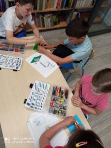 Na zdjęciu znajdują się uczestnicy warsztatów. Dzieci wykonają konstrukcje z klocków lego - siedzą na krzesłach. Zdjęcie wykonano w bibliotece.