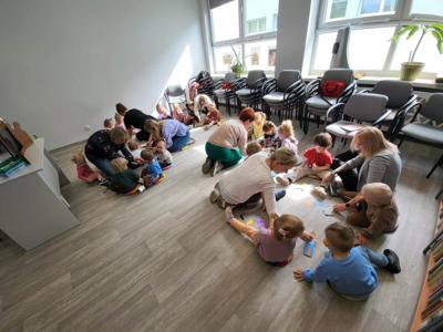 Na zdjęciu znajdują się uczestnicy warsztatów. Dzieci siedzą na poduszkach i wykonują prace plastyczne. Zdjęcie wykonano w bibliotece.