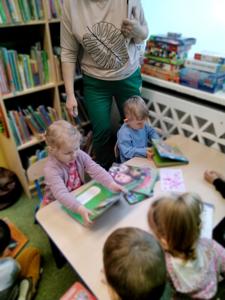 Na zdjęciu znajdują się uczestnicy warsztatów. Dzieci siedzą przy stole i oglądają książki. Zdjęcie wykonano w bibliotece.