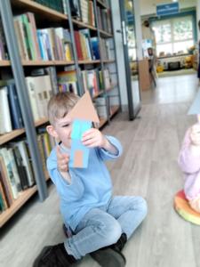 Na zdjęciu znajdują się uczestnicy warsztatów. Dzieci siedzą na poduszkach i wykonują prace plastyczne. Zdjęcie wykonano w bibliotece.