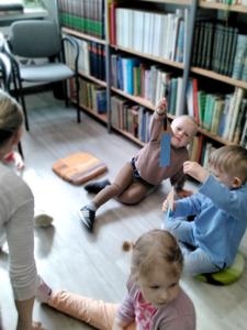 Na zdjęciu znajdują się uczestnicy warsztatów. Dzieci siedzą na poduszkach i wykonują prace plastyczne. Zdjęcie wykonano w bibliotece.