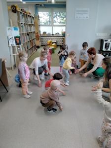 Na zdjęciu znajdują się uczestnicy warsztatów. Dzieci stoją w kręgu i wykonują zadania. Zdjęcie wykonano w bibliotece.