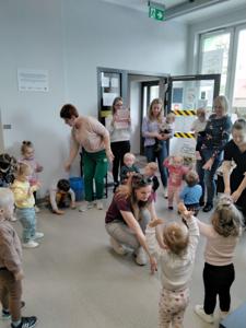 Na zdjęciu znajdują się uczestnicy warsztatów. Dzieci stoją w kręgu i wykonują zadania. Zdjęcie wykonano w bibliotece.