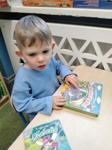 Na zdjęciu znajdują się uczestnicy warsztatów. Dzieci oglądają książki. Zdjęcie wykonano w bibliotece.