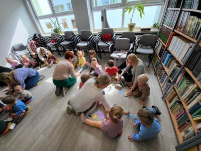 Na zdjęciu znajdują się uczestnicy warsztatów. Dzieci siedzą na poduszkach i wykonują prace plastyczne. Zdjęcie wykonano w bibliotece.