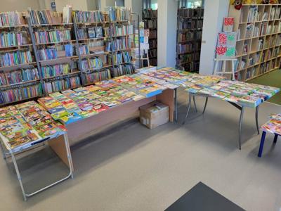 Na zdjęciu znajdują książki poukładane na stołach. W tle księgozbiór biblioteki.