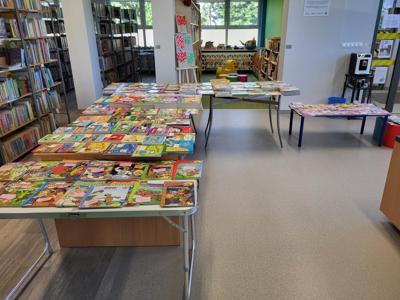 Na zdjęciu znajdują książki poukładane na stołach. W tle księgozbiór biblioteki.