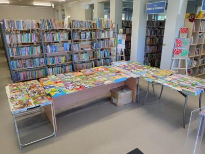 Na zdjęciu znajdują książki poukładane na stołach. W tle księgozbiór biblioteki.