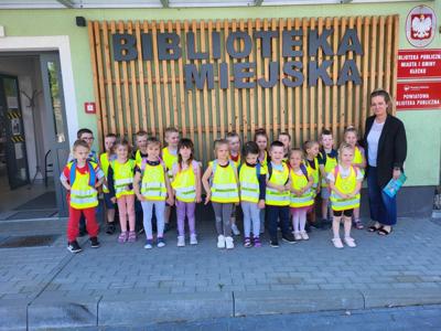 Na zdjęciu znajdują się uczestnicy kiermaszu. Dzieci stoją. Zdjęcie wykonano przed wejściem do biblioteki. w tle napisz Biblioteka Miejska.