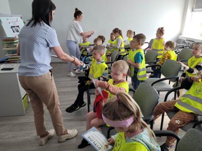 Na zdjęciu znajdują się uczestnicy zajęć. Dzieci siedzą na krzesłach i oglądają książki. Zdjęcie wykonano w bibliotece.
