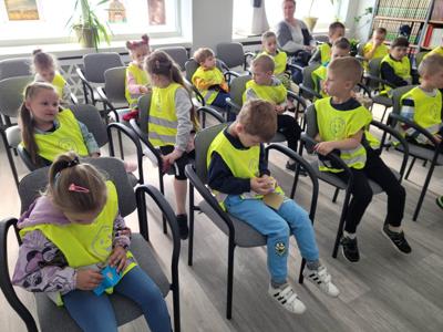 Na zdjęciu znajdują się uczestnicy zajęć. Dzieci siedzą na krzesłach i oglądają książki. Zdjęcie wykonano w bibliotece.