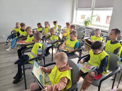 Na zdjęciu znajdują się uczestnicy zajęć. Dzieci siedzą na krzesłach i oglądają książki. Zdjęcie wykonano w bibliotece.