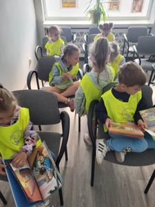 Na zdjęciu znajdują się uczestnicy zajęć. Dzieci siedzą na krzesłach i oglądają książki. Zdjęcie wykonano w bibliotece.