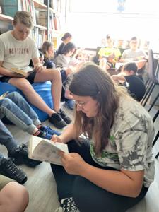 Na zdjęciu znajduje się młodzież czytająca książki. Młodzież siedzi - w tle księgozbiór biblioteki.