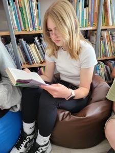 Na zdjęciu znajduje się młodzież czytająca książki. Młodzież siedzi - w tle księgozbiór biblioteki.