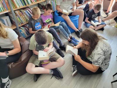Na zdjęciu znajduje się młodzież czytająca książki. Młodzież siedzi - w tle księgozbiór biblioteki.
