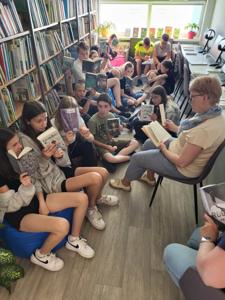 Na zdjęciu znajduje się młodzież czytająca książki. Młodzież siedzi - w tle księgozbiór biblioteki.