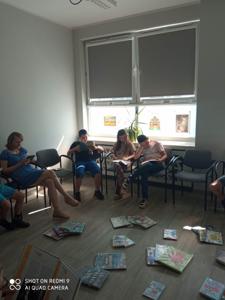 Na zdjęciu znajduje się młodzież czytająca książki. Młodzież siedzi - w tle księgozbiór biblioteki.