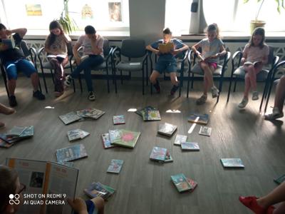 Na zdjęciu znajduje się młodzież czytająca książki. Młodzież siedzi - w tle księgozbiór biblioteki.