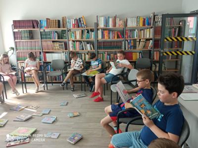 Na zdjęciu znajduje się młodzież czytająca książki. Młodzież siedzi - w tle księgozbiór biblioteki.