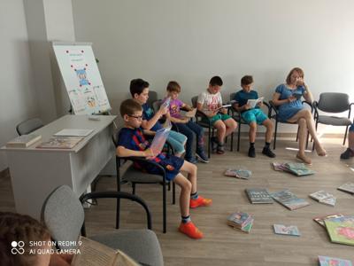 Na zdjęciu znajduje się młodzież czytająca książki. Młodzież siedzi - w tle księgozbiór biblioteki.