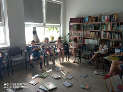 Na zdjęciu znajduje się młodzież czytająca książki. Młodzież siedzi - w tle księgozbiór biblioteki.