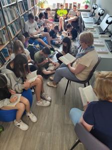 Na zdjęciu znajduje się młodzież czytająca książki. Młodzież siedzi - w tle księgozbiór biblioteki.