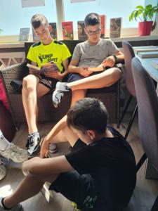 Na zdjęciu znajduje się młodzież czytająca książki. Młodzież siedzi - w tle księgozbiór biblioteki.