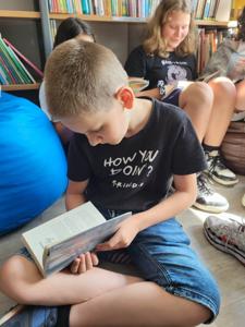 Na zdjęciu znajduje się młodzież czytająca książki. Młodzież siedzi - w tle księgozbiór biblioteki.