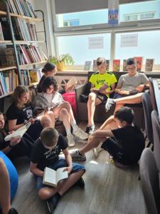 Na zdjęciu znajduje się młodzież czytająca książki. Młodzież siedzi - w tle księgozbiór biblioteki.