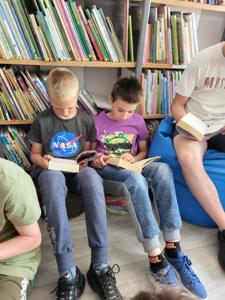 Na zdjęciu znajduje się młodzież czytająca książki. Młodzież siedzi - w tle księgozbiór biblioteki.